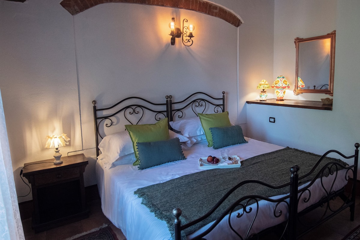 A neatly made bed with a green throw blanket, a bedside table with a lamp, and a wall-mounted mirror.