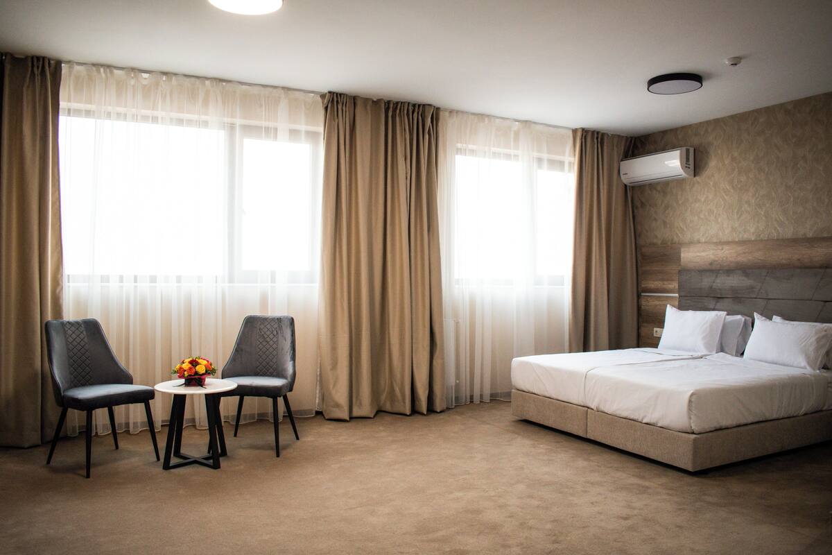 A hotel room with a bed, two chairs, a small table with a flower vase, and a large window with curtains.