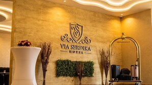 Un espace d’accueil avec une enseigne fixée au mur indiquant « Via Serdika Hotel », un comptoir de réception blanc, une plante verte fixée au mur et un chariot à bagages.