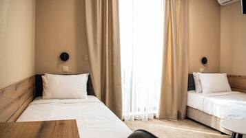 A hotel room with two single beds, a wooden headboard, a small round table, and a window with sheer curtains.