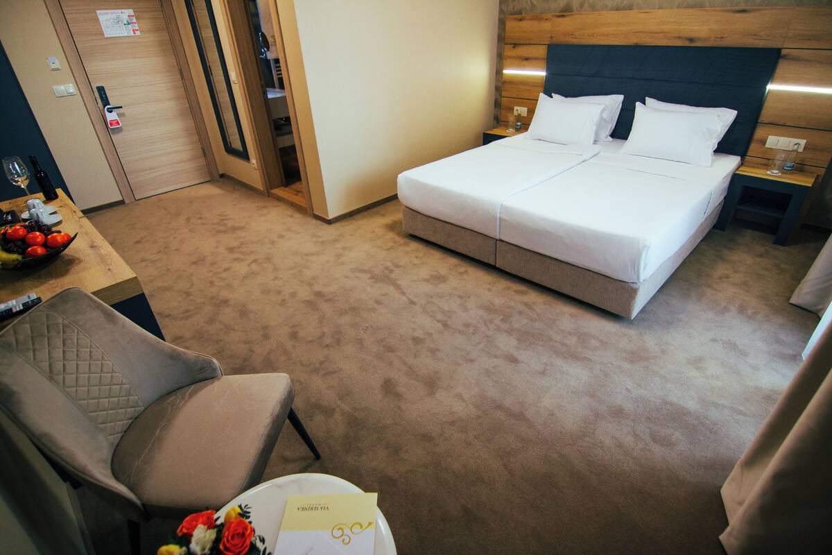 A hotel room with a bed, a chair, a small table with a bowl of fruit, and a nightstand with a lamp.