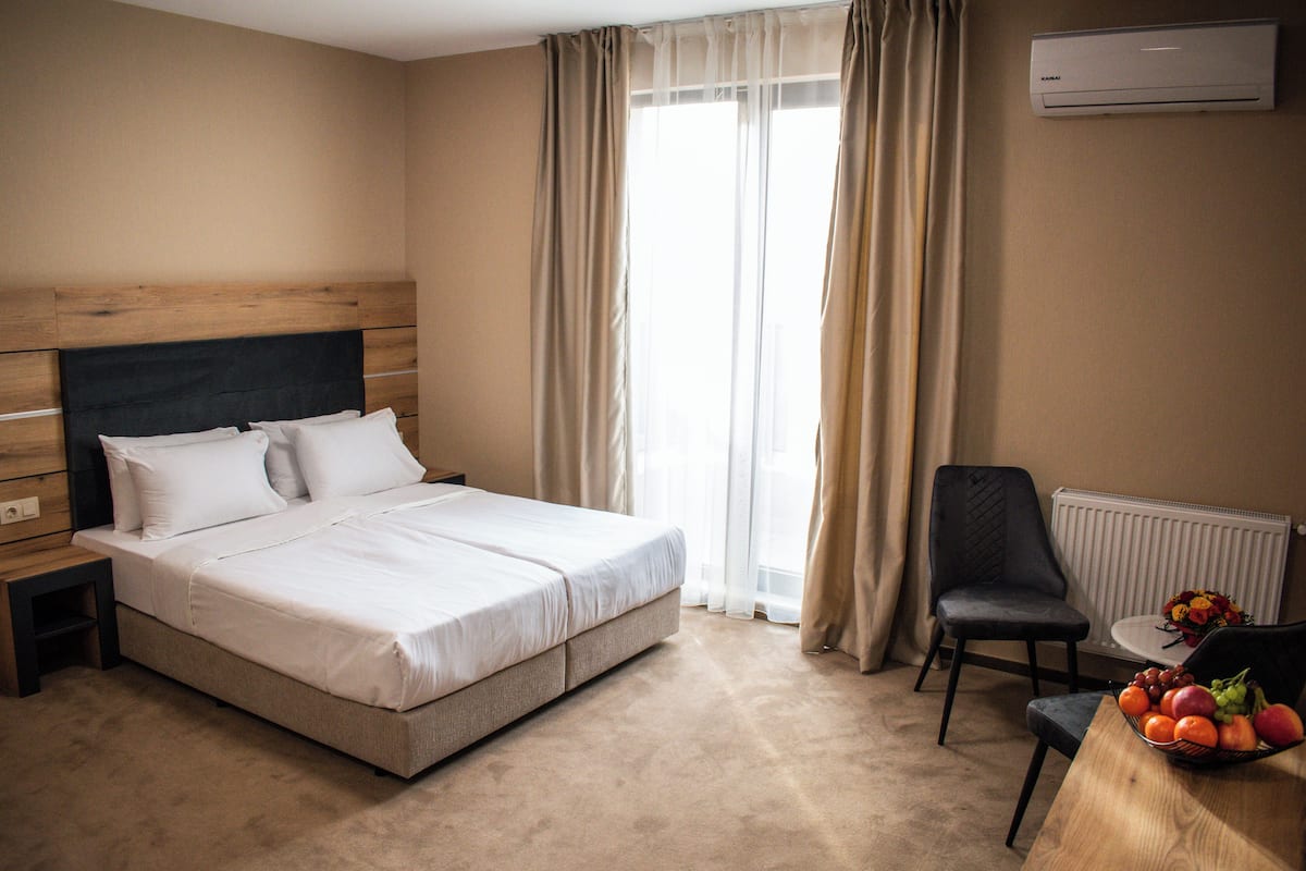 A hotel room with a bed, a chair, a table with fruit, and an air conditioning unit.