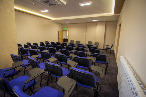 A conference room with rows of blue chairs and black tables, a white radiator, and a door with an exit sign. - Hotel Via Serdika (Sofia)