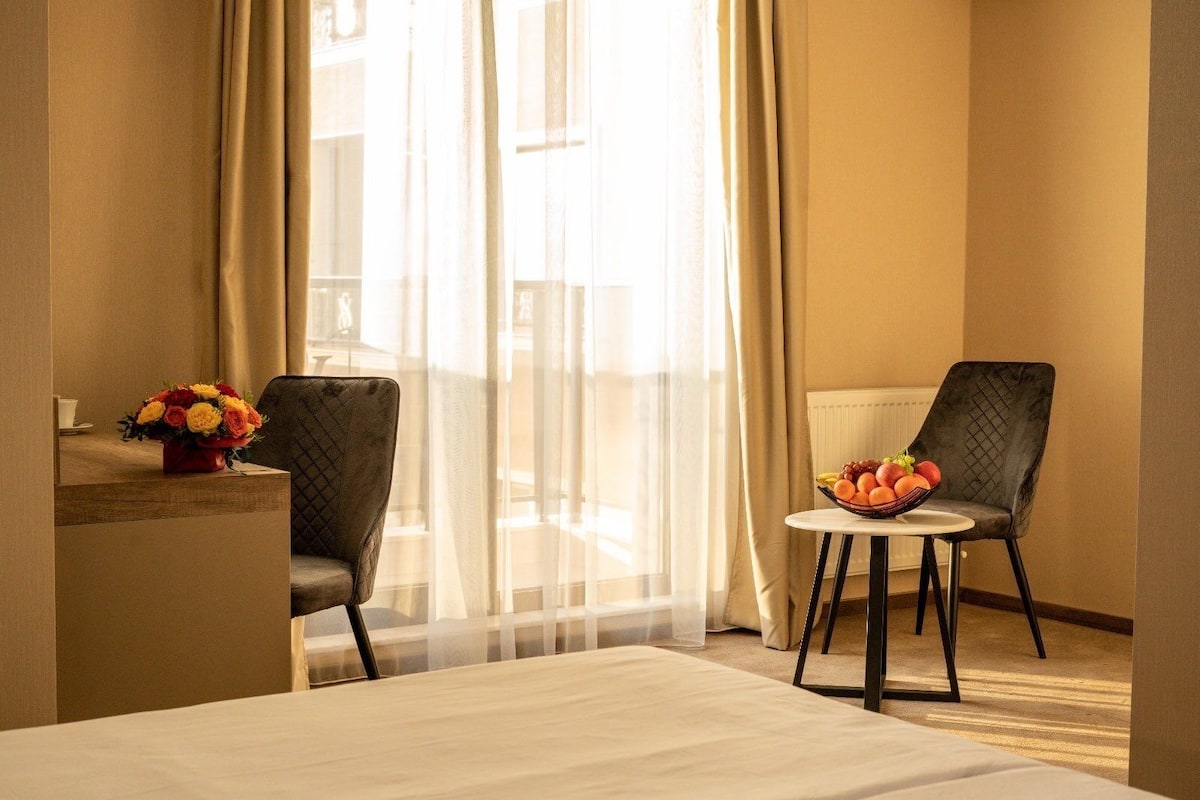 A hotel room with a bed, two armchairs, a small table with fruit, and a vase of flowers.