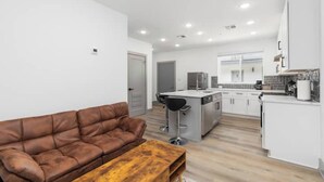 Un apartamento moderno con un sofá de cuero marrón, una mesa de centro de madera, una cocina con armarios blancos y una pequeña isla con un lavamanos.