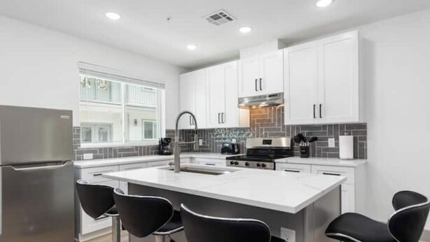 Una cocina moderna con armarios blancos, un refrigerador de acero inoxidable, una isla central con taburetes altos y un ventanal que proporciona luz natural.