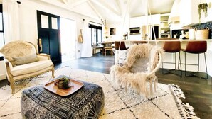 A spacious living room with a high ceiling, a cozy armchair, a patterned ottoman, and a fluffy dog.