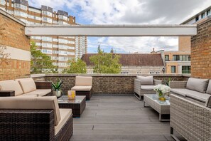 Eine Dachterrasse mit Korbmöbeln, einem Glastisch und Blick auf die umliegenden Gebäude.