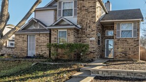 A two-story brick house with a front porch, a small garden, and a visible address number. - Private Backyard + Deck - Near DFW + Toyota Music (Euless)
