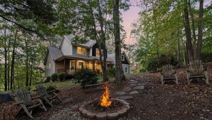 A house with a fire pit in the yard, surrounded by trees and wooden chairs.