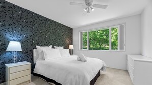 Un dormitorio con cama, mesitas de noche, ventilador de techo y ventana con vista a árboles.
