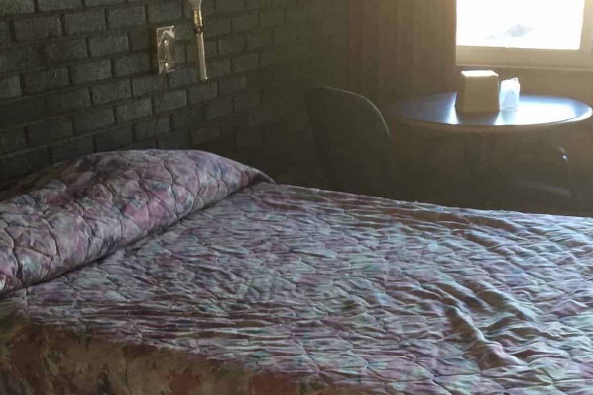 A room with a bed, a brick wall, a window, and a small table with a lamp.