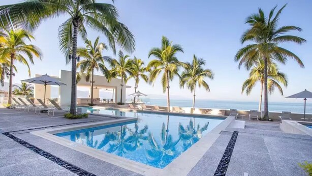 A swimming pool with lounge chairs and palm trees, overlooking the ocean. - Luxury Oceanfront Condo with WiFi, Pool, and Fitness Room in Puerto Vallarta (Puerto Vallarta)