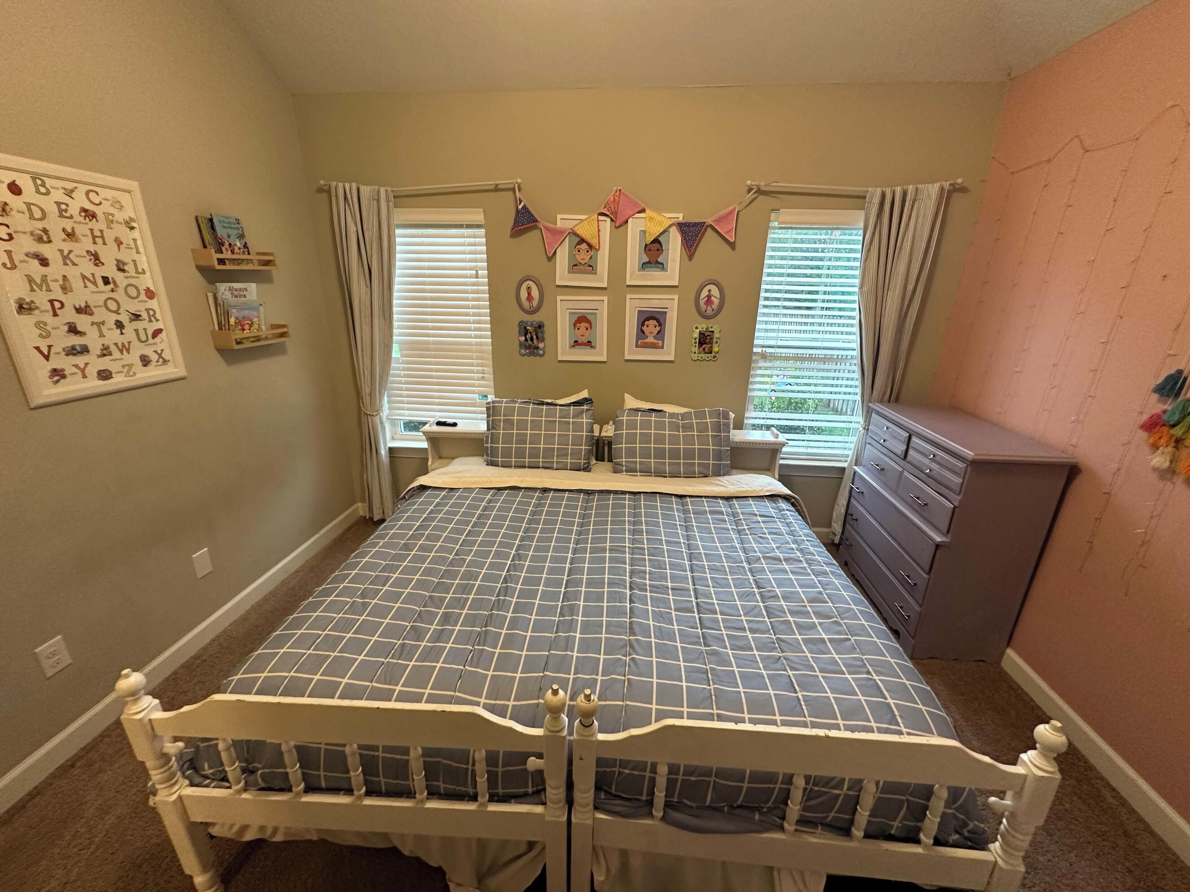 A bedroom with a bed, a dresser, a window with blinds, a wall with framed pictures, and a shelf with books.