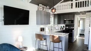 Un vaste espace de vie comprenant un grand téléviseur, un ventilateur de plafond et un coin bar avec des tabourets.