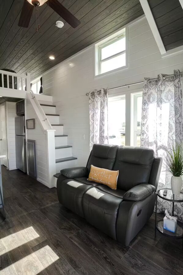 A spacious living area with a black leather sofa, a staircase, a kitchen, and large windows.