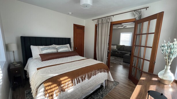 A bedroom with a large bed, a nightstand, a bedside lamp, a wooden floor, a ceiling fan, and a view into the living area through an open door.