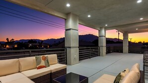 An outdoor seating area with cushioned sofas and a glass coffee table, overlooking a sunset with mountains in the background. - Spacious New Build - Heated Pool, Covered Patio, Sunsets, Games, Luxurious Space (Desert Hot Springs)