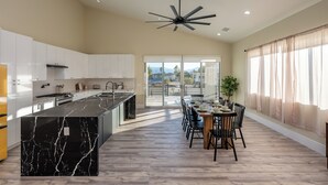 A modern kitchen with a marble island, a dining area with chairs, and a ceiling fan. - Spacious New Build - Heated Pool, Covered Patio, Sunsets, Games, Luxurious Space (Desert Hot Springs)