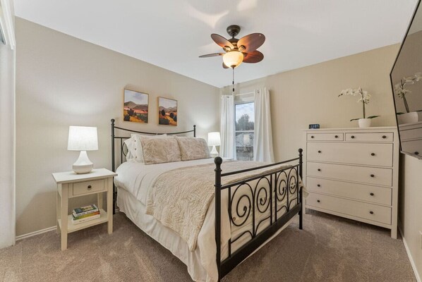 A bedroom with a bed, bedside tables, a dresser, a ceiling fan, and artwork on the walls.