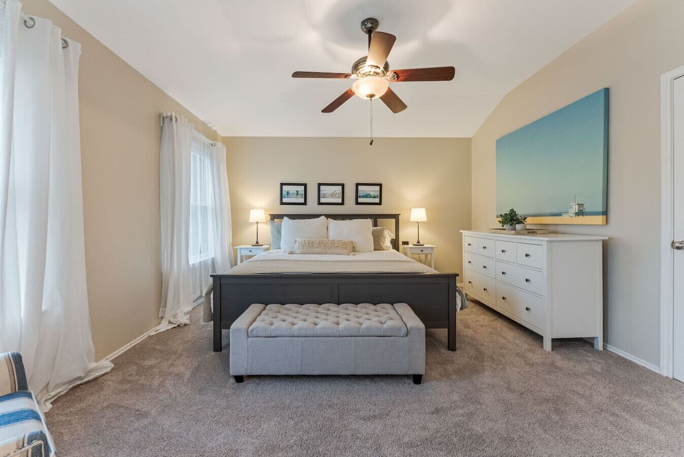 Une chambre à coucher comprenant un lit, un ventilateur de plafond, une table de chevet, une commode et un grand tableau accroché au mur.