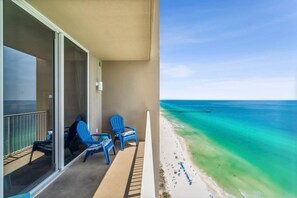 Ein Balkon mit zwei blauen Stühlen und einem Tisch, der auf einen Strand mit klarem türkisblauem Wasser und einem Steg blickt.