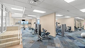 A spacious gym with various exercise equipment, a patterned carpet, and large windows providing natural light. - Snowbirds Land on the 29th Floor for the Winter (Panama City Beach)