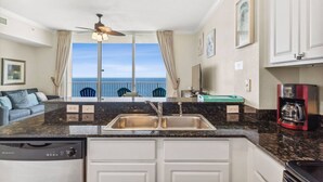 A kitchen with a granite countertop, a dishwasher, and a coffee maker. There is a view of the ocean through large windows. - Snowbirds Land on the 29th Floor for the Winter (Panama City Beach)