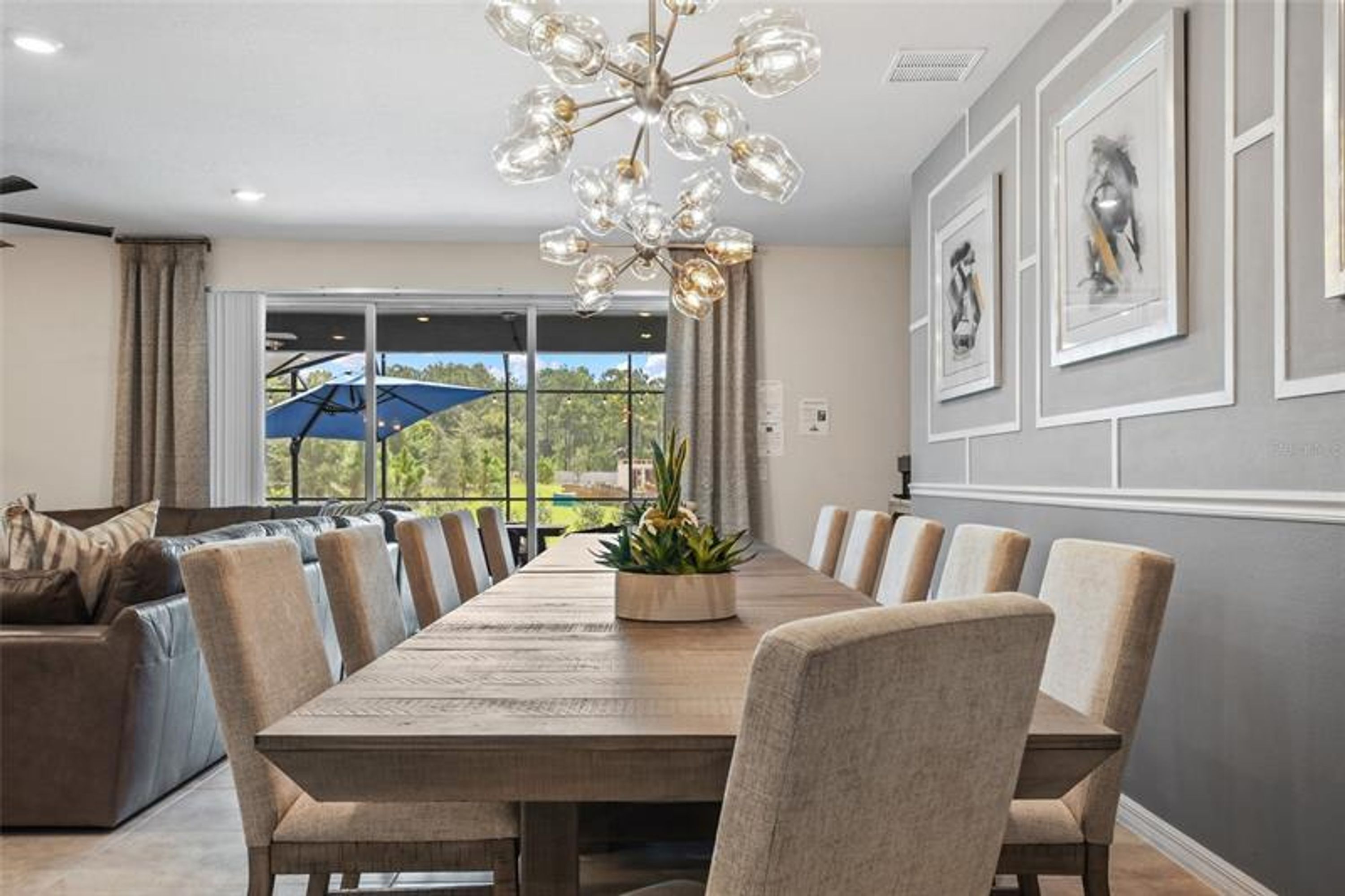 Ein Essbereich mit einem langen Tisch, Stühlen und einer Chandelier. Ein Sofa und ein Fenster mit Blick auf Grünflächen.