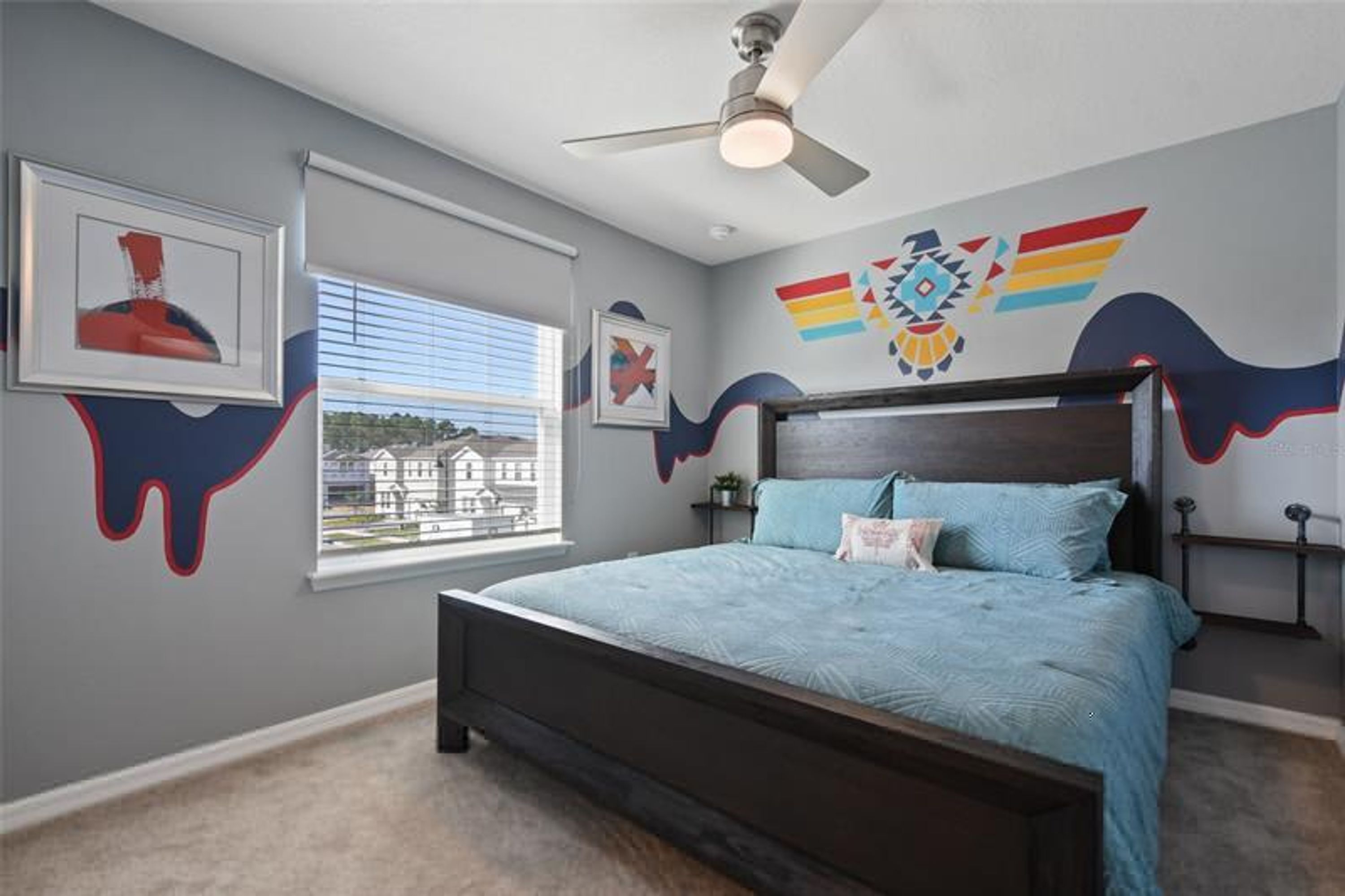 Une chambre à coucher avec un lit, un ventilateur de plafond, une fenêtre avec des stores et un mur orné d’un motif coloré.