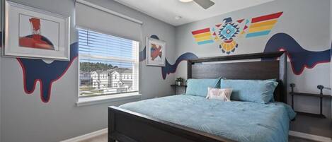 Une chambre à coucher avec un lit, un ventilateur de plafond, une fenêtre avec des stores et un mur orné d’un motif coloré.