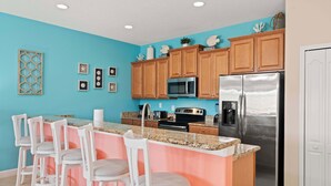 Une cuisine aux murs de couleur turquoise, avec des meubles en bois, un réfrigérateur en acier inoxydable et un coin bar doté de tabourets blancs.