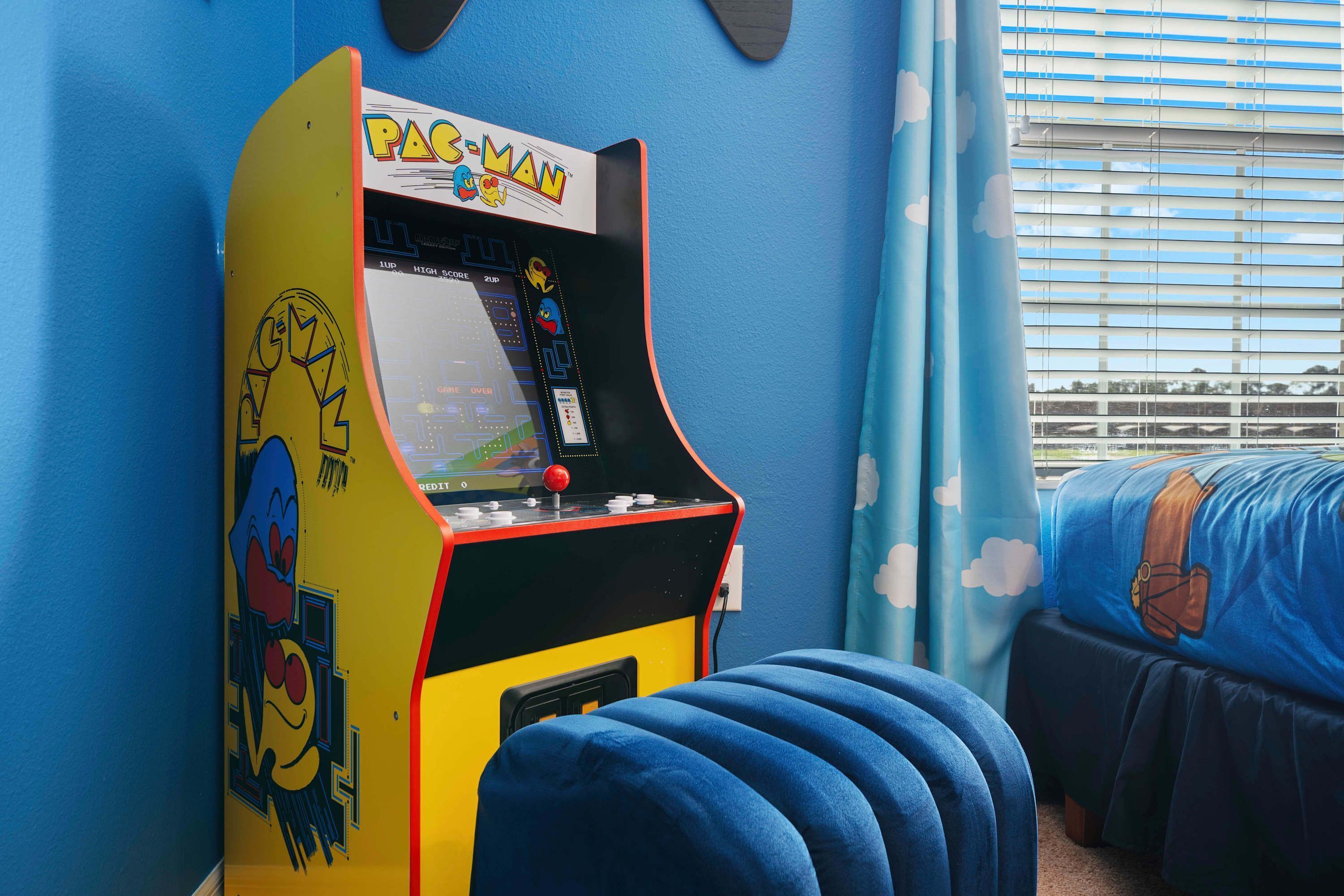 Une borne d’arcade Pac-Man dans une pièce avec un mur bleu et un lit recouvert d’une couette aux motifs de dessins animés.