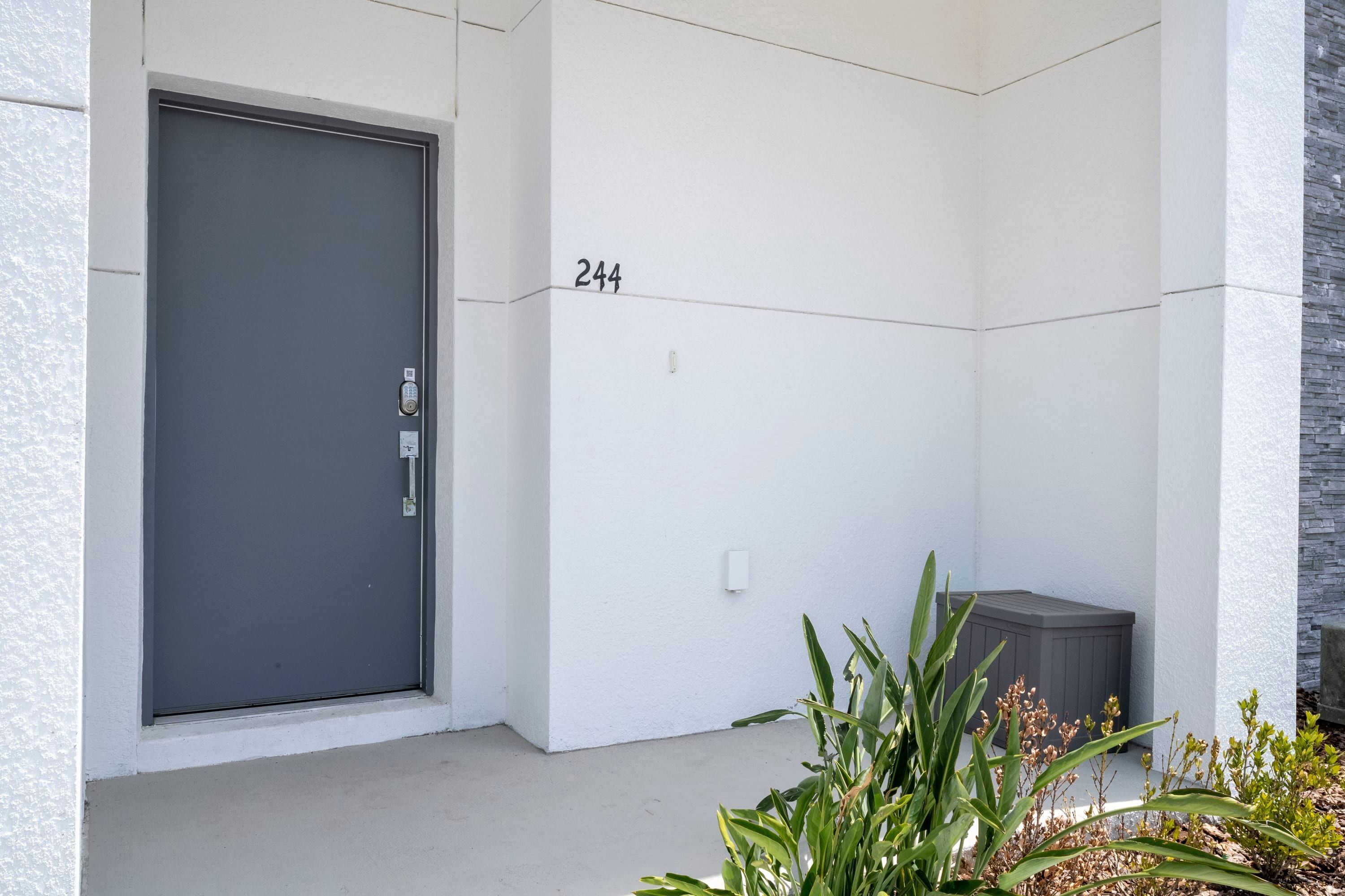 Une entrée d’immeuble moderne avec une porte grise, un mur blanc portant le numéro 244 et une poubelle grise.