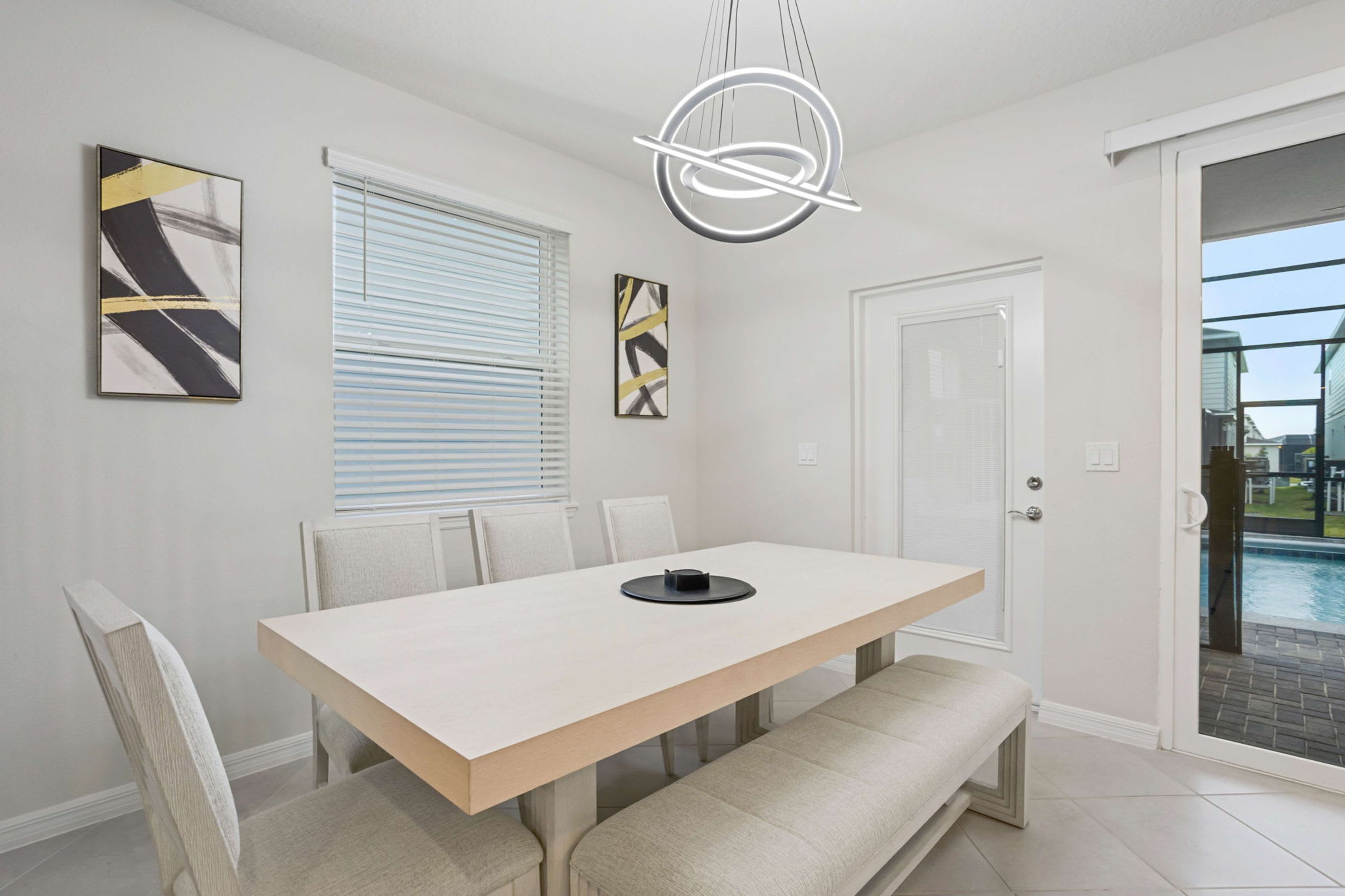 A modern dining area with a rectangular table, six chairs, a bench, abstract wall art, a contemporary light fixture, and a sliding glass door leading to an outdoor pool area.