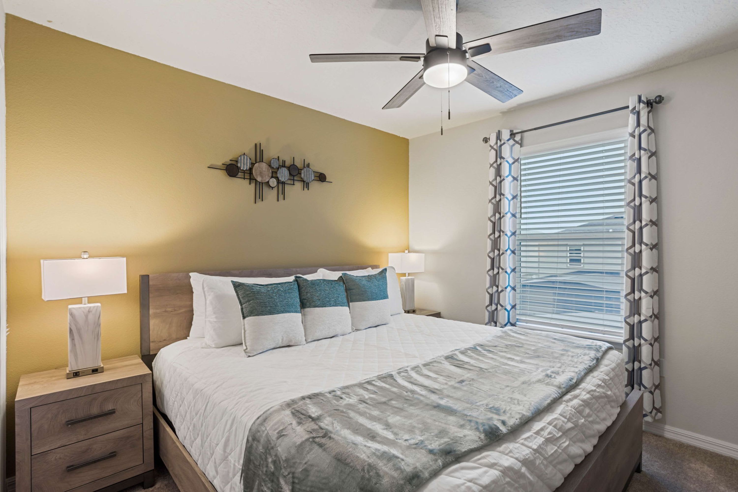 Quarto de hotel com uma cama, duas mesas de cabeceira com luminárias, um ventilador de teto e uma janela com cortinas estampadas.