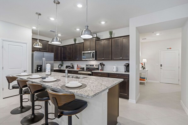 A modern kitchen with dark wood cabinets, a granite countertop, stainless steel appliances, and pendant lighting. - Brand New 5 bed Villa in Windsor Cay (Clermont)