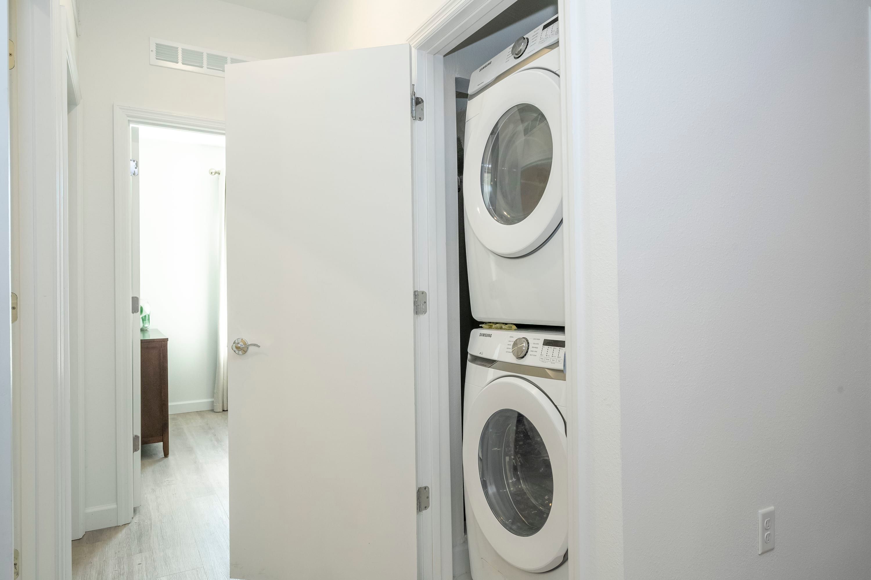 A laundry room with two stacked washing machines, a door leading to another room, and a small table with a green object on it.