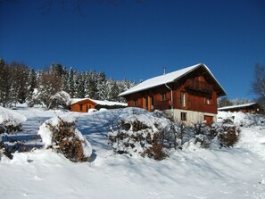 A wooden cabin with a snow-covered roof, surrounded by snow-covered trees and bushes.