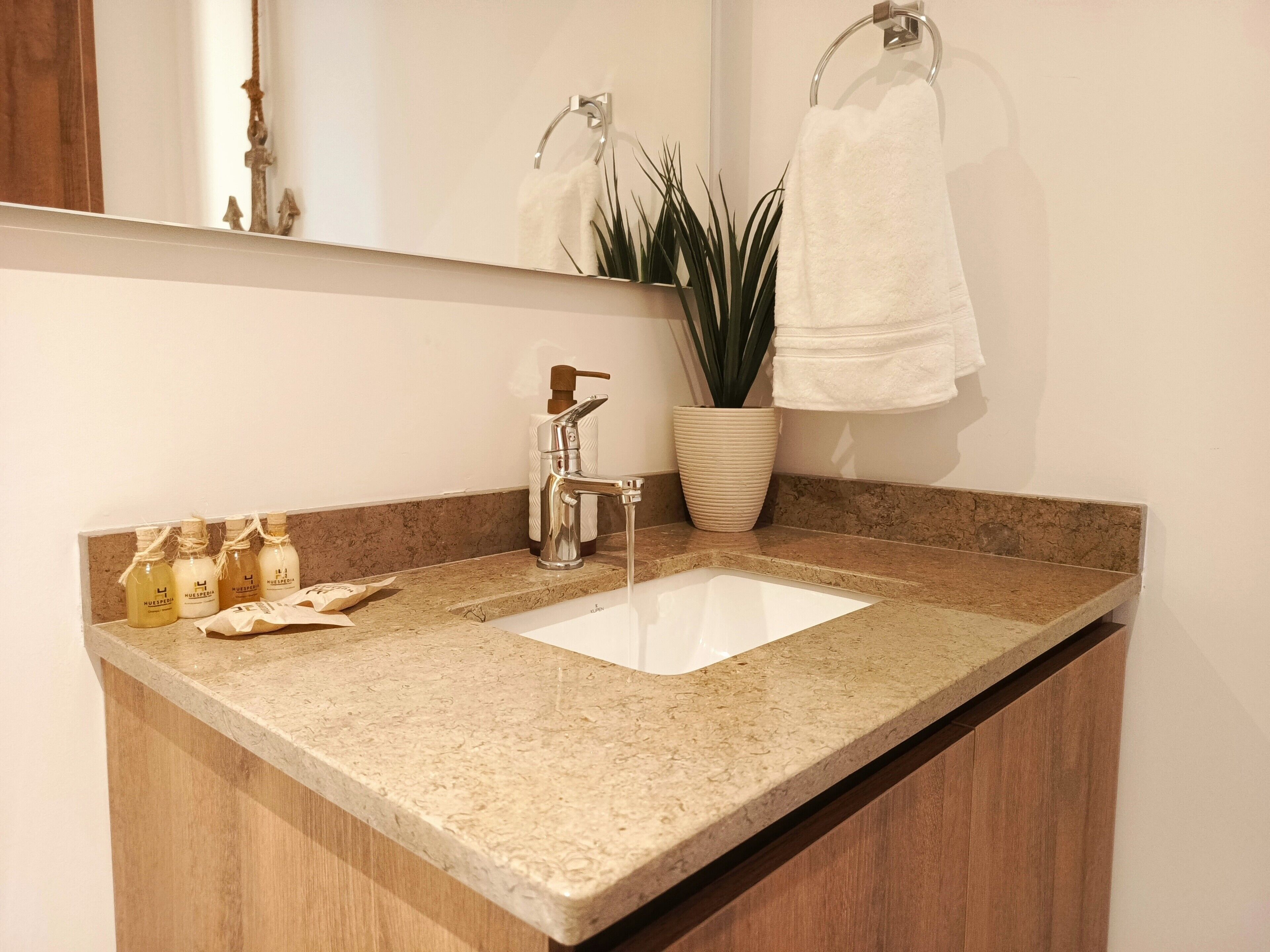 A bathroom with a granite countertop, a white sink, a mirror, a towel rack, a potted plant, and toiletries.