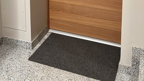 Eine Holztür mit digitalem Schloss und Schlüsselloch, eine kleine Fußmatte im Eingangsbereich und ein an der Wand montiertes Bedienfeld.