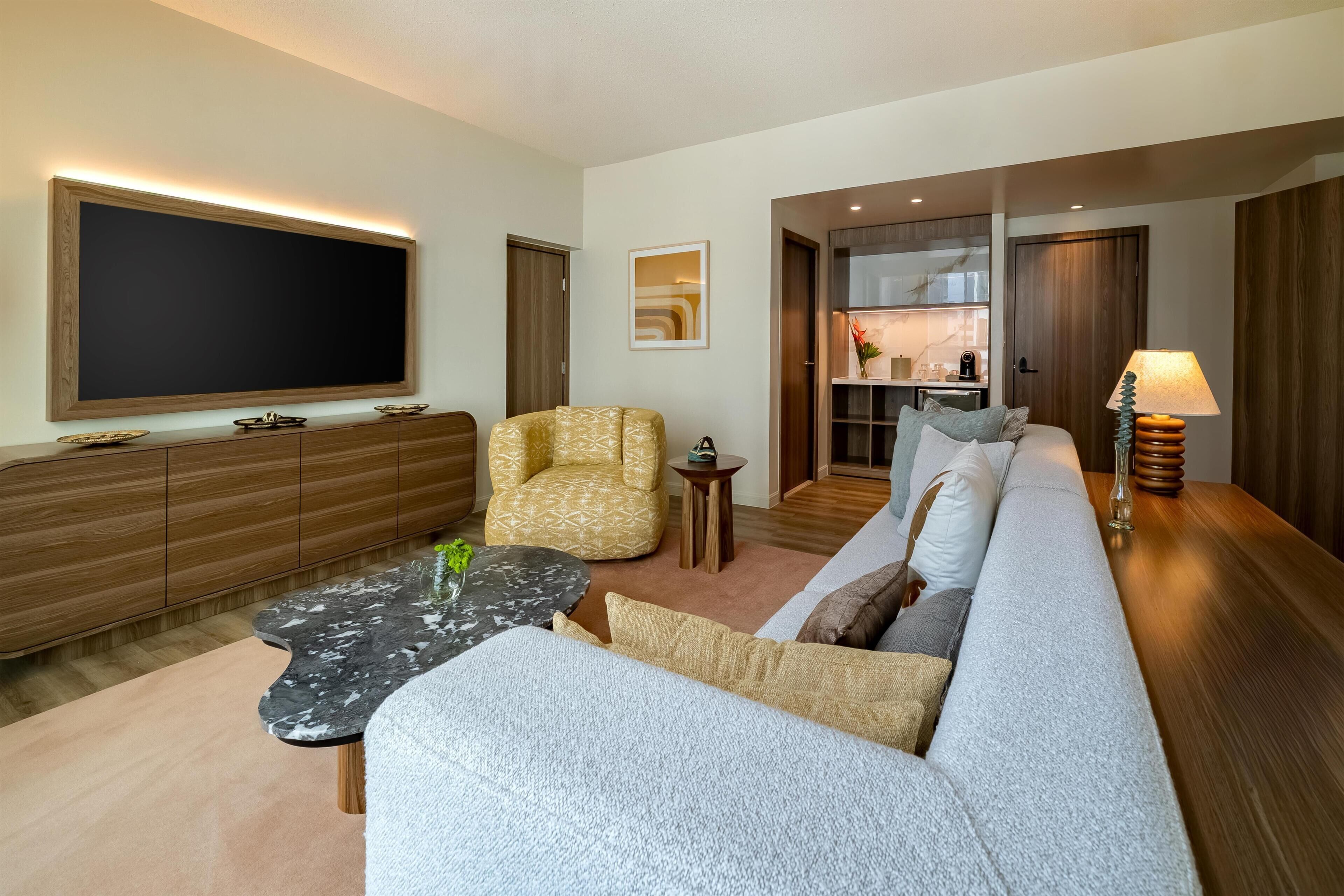 A modern living room with a large flat-screen TV, a sofa, a patterned armchair, a coffee table, and a lamp.