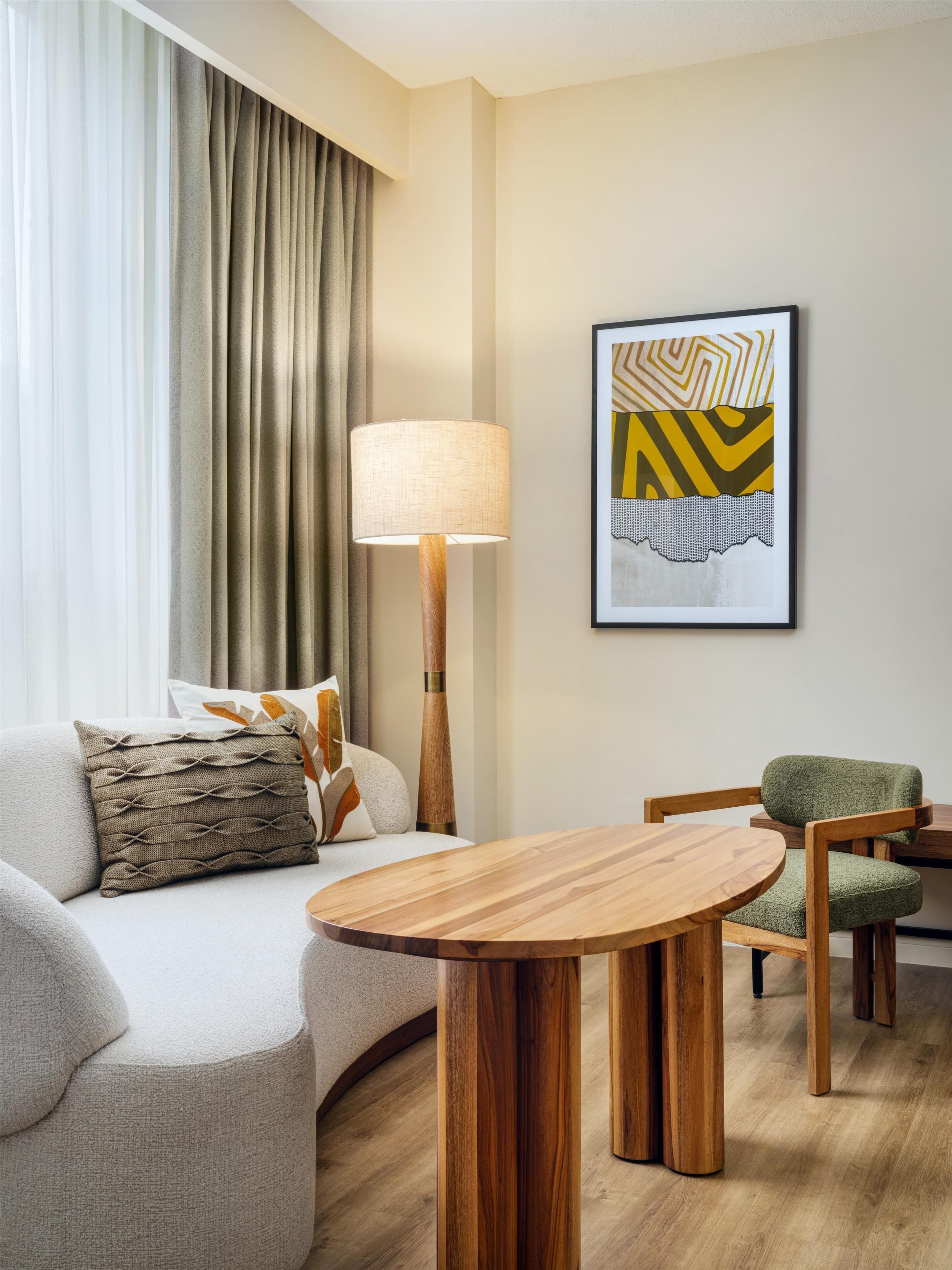 A modern hotel room with a sofa, a round wooden table, a lamp, and a framed abstract artwork on the wall.