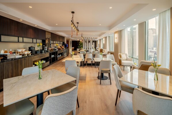 Un área de comedor de hotel moderna con una cocina al fondo, ventanas amplias y muebles contemporáneos.