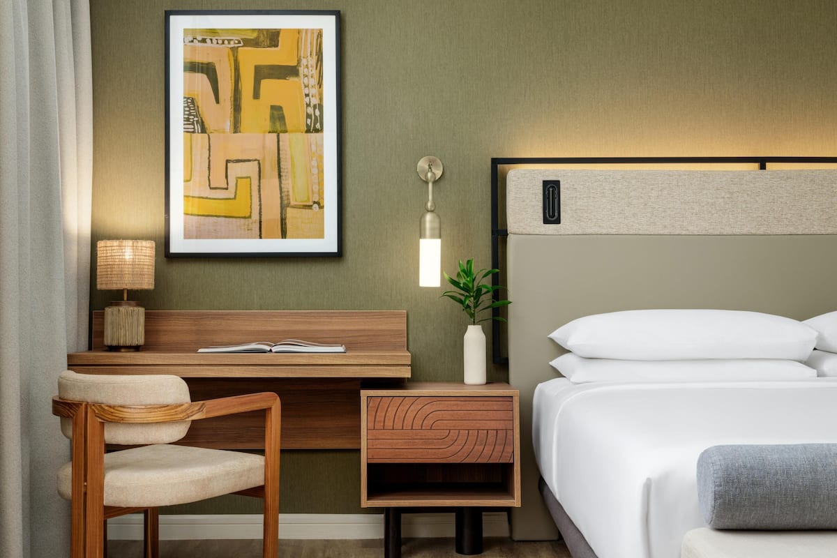A hotel room with a bed, a desk with a chair, a nightstand, a lamp, a framed artwork, and a potted plant.