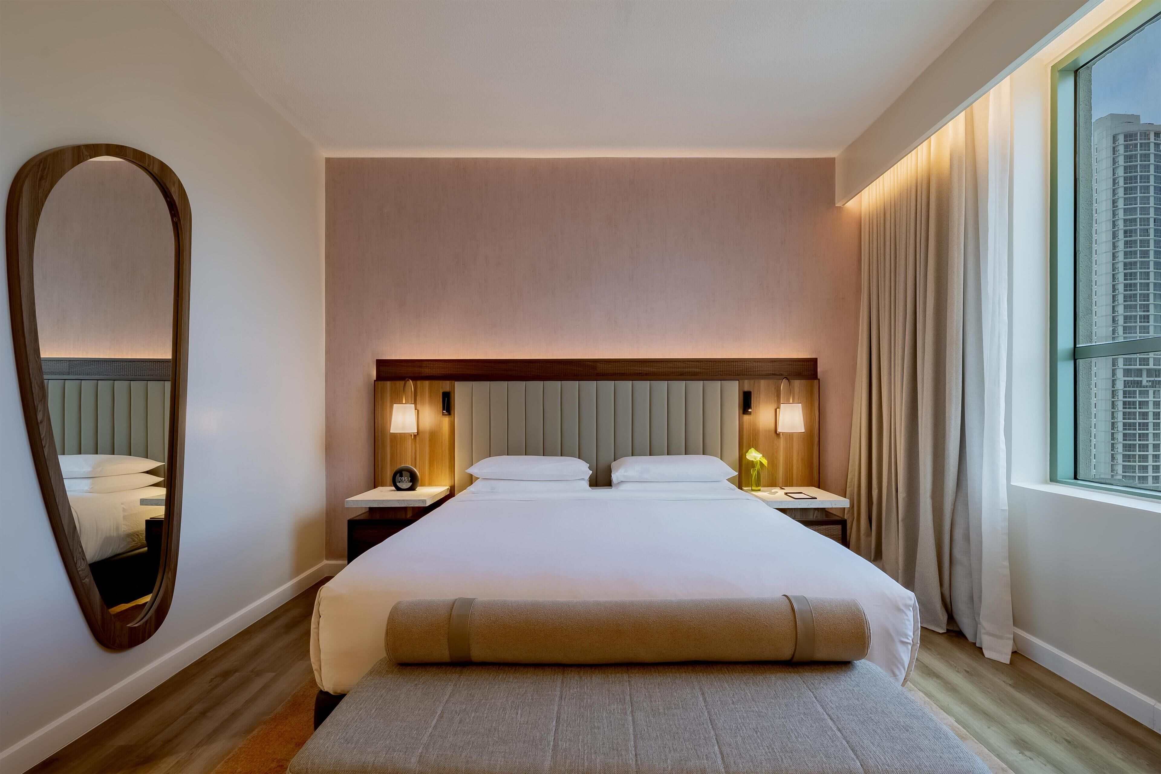 Une chambre d’hôtel avec un grand lit, des lampes de chevet, un miroir et une fenêtre avec des rideaux.