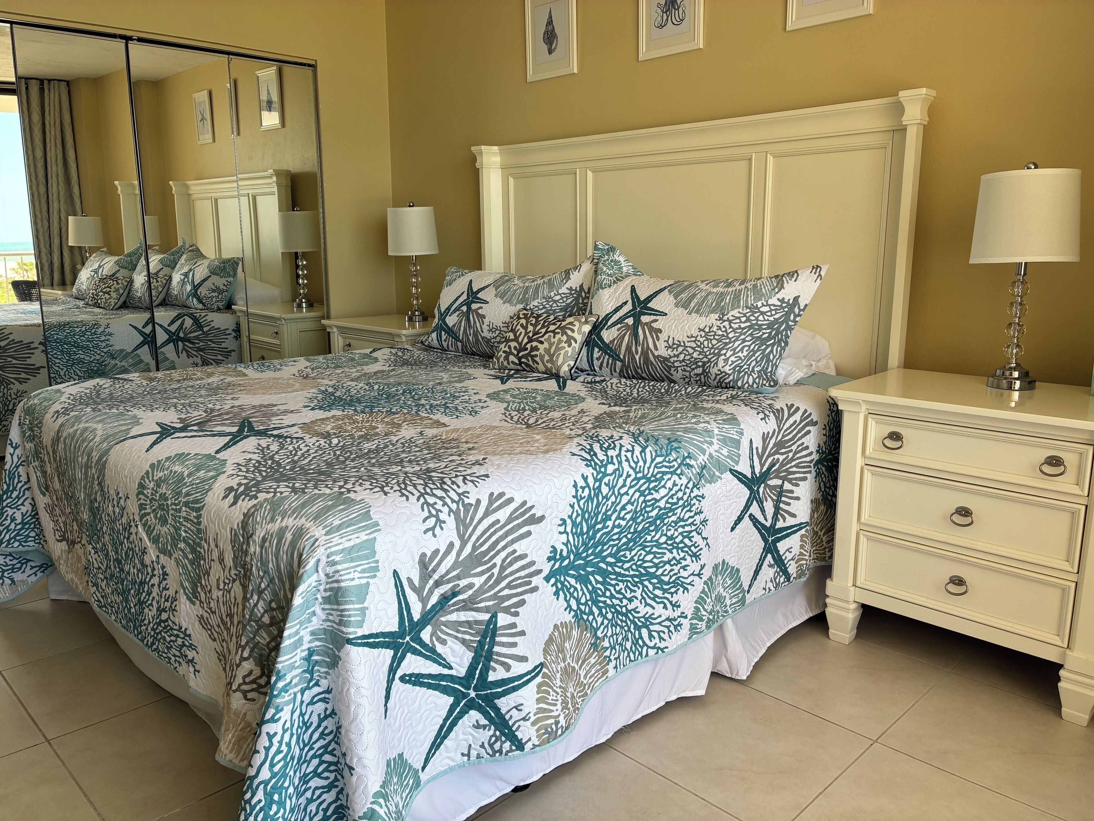 Un dormitorio con una cama que presenta un edredón estampado con motivos tropicales, una mesita de noche con una lámpara y un espejo grande en la pared.