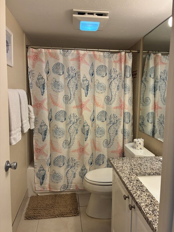 A bathroom with a shower curtain featuring a seahorse and shell pattern, a toilet, a sink with a granite countertop, and a small rug on the floor.