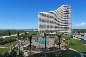 A modern hotel with a curved facade, a central swimming pool, and a well-maintained lawn with palm trees.
