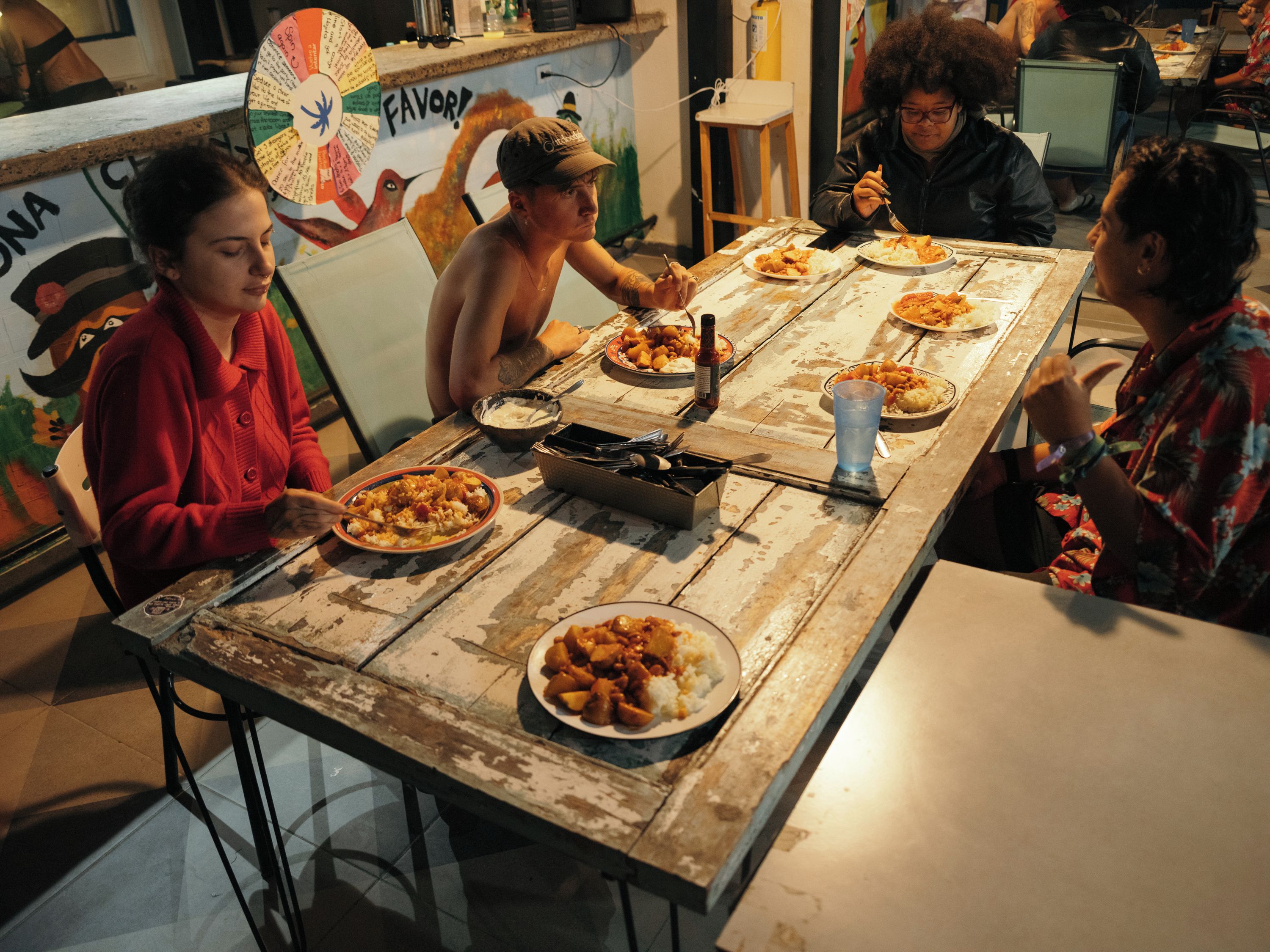 Un grupo de personas cenando en una mesa de madera rústica, con varios platos y un abanico al fondo.